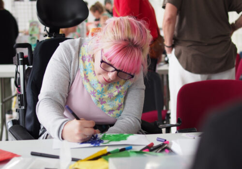 A young woman with pink hair is using watercolour pens to create a work of art.
