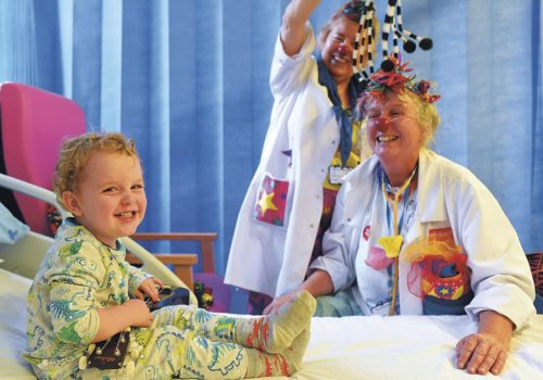 Two Clown Doctors are engaging a young child on a hospital ward in creative play. All three are laughing.