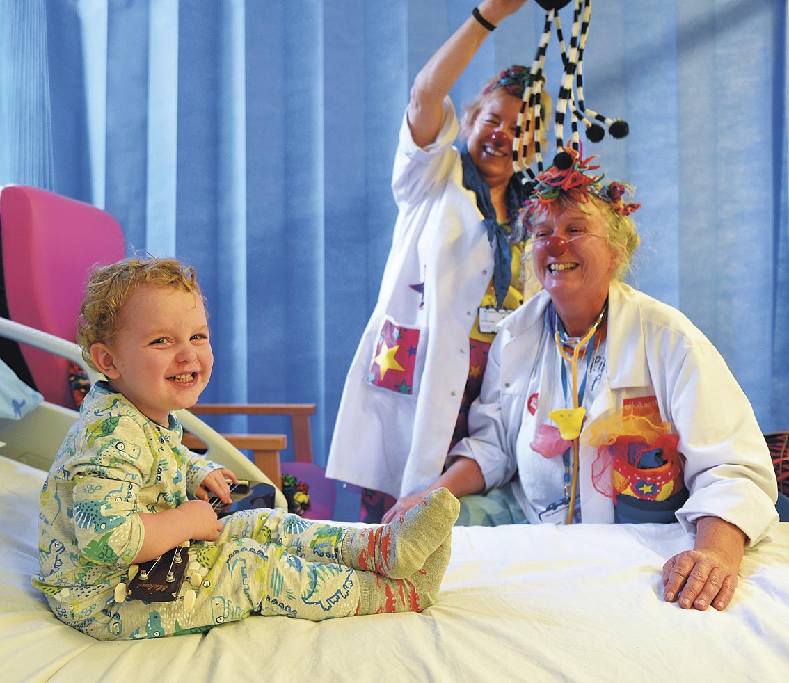 Two Clown Doctors are engaging a young child on a hospital ward in creative play. All three are laughing.