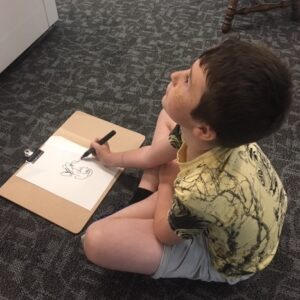 The image shows a young boy staring intently at an object that is out of view whilst drawing it in black ink on a piece of paper in front of him.