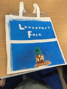 A tote bag in brilliant blue acrylic paint with a hand coloured picture of a beach hut under the words Lowestoft Folk