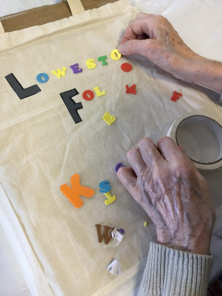 The image shows a pair of hands, obviously of an elderly person, delicately placing a sticky-backed letter on to a bag as she spells out the word Lowestoft