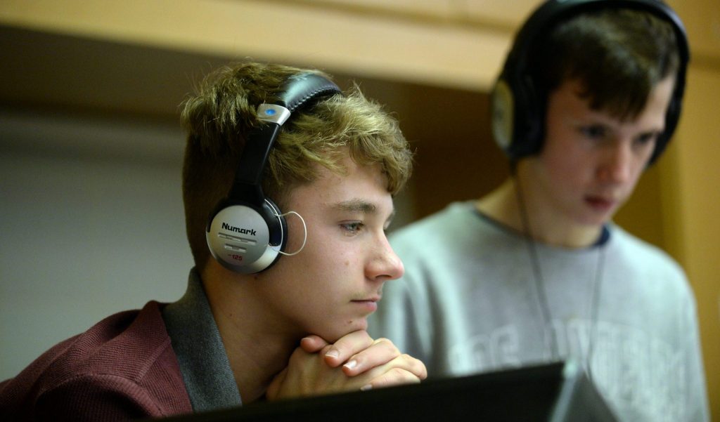 In the foreground, a young man with headphones stares intently at the DJ screen, whilst his partner stands behind, also watching the DJ decks