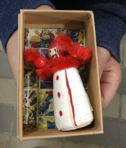 A small wooden figure, decorated in red and white, inside a tiny cardboard box, decorated with superhero paper
