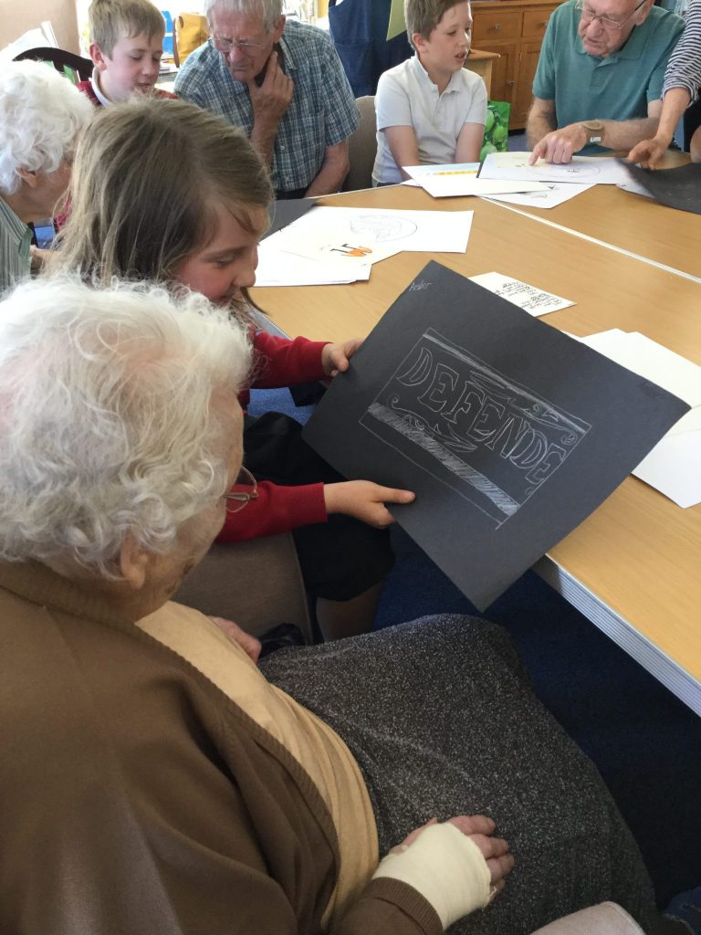 A young person is showing an elderly lady a picture, drawn in white pencil on black paper, whilst in the background other children can be seen, talking abut the pictures they have drawn