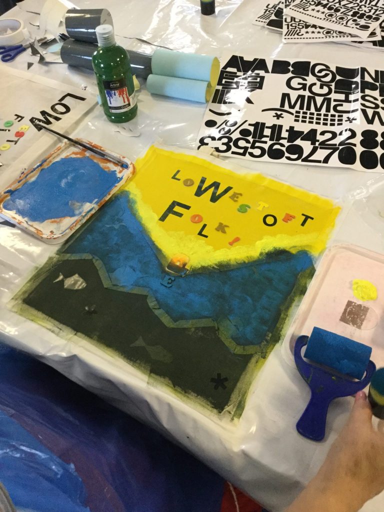 A decorated canvas bag, the top half bright yellow with the words Lowestoft Folk clearly visible and the bottom half a deep blue descending into black at the bottom of the picture. Around the bag there are sheets of sticky-backed letters, rollers and a bottle of green acrylic paint