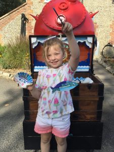 A young girl stands in front of the Sea Chest holding up a scallop shell she has decorated along with the fish she made
