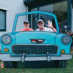 2 adults posing behind a 2D blue car front