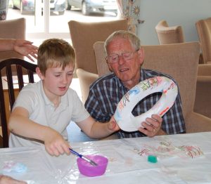 A young boy dipping a brush in a pot of glue whilst the man beside him is holding a polystyrene ring onto which they are pasting pieces of decorated paper