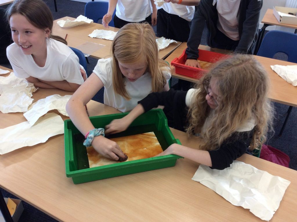 Three girls are seated at a school table, two of them are holding a piece of paper in a green tray full of dark liquid it's cold tea - and are rubbing the paper with the tea bag