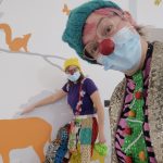 Clown doctor in foreground and clown doctor in background in front of a wall with plants painted in grey and animals painted in orange
