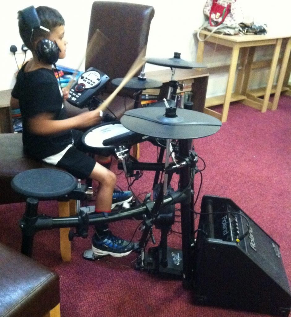 A young boy seated at an electric drum kit drumming so fast the sticks are just a blur