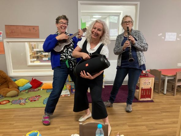 Three smiling musicians who are dancing with their instruments and a speaker. One musician is playing a ukulele, one a clarinet and the third is holding a boombox. Around them are toys and mats on the floor in readiness for an early years children's music activity.