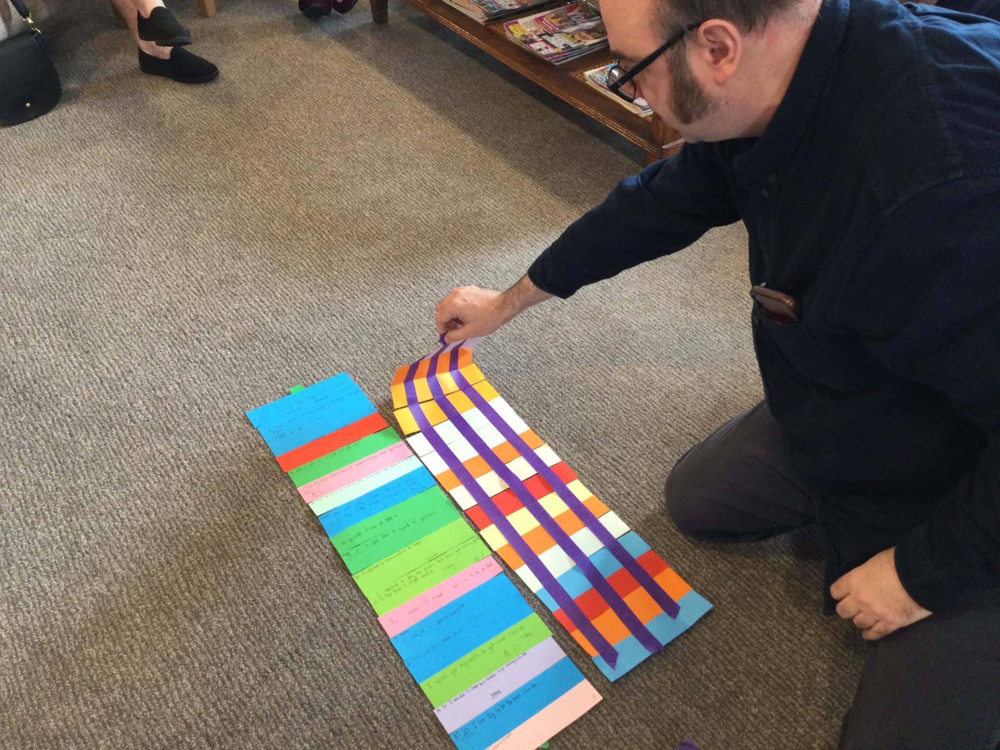 A man kneeling on the carpeted floor, with strips of coloured paper laid out before him