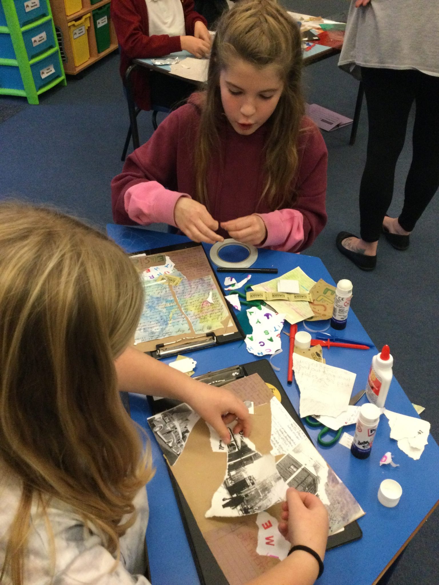 Two children seated at a table with glue, tape and an assortment of pictures which they are tearing up to use in their collage