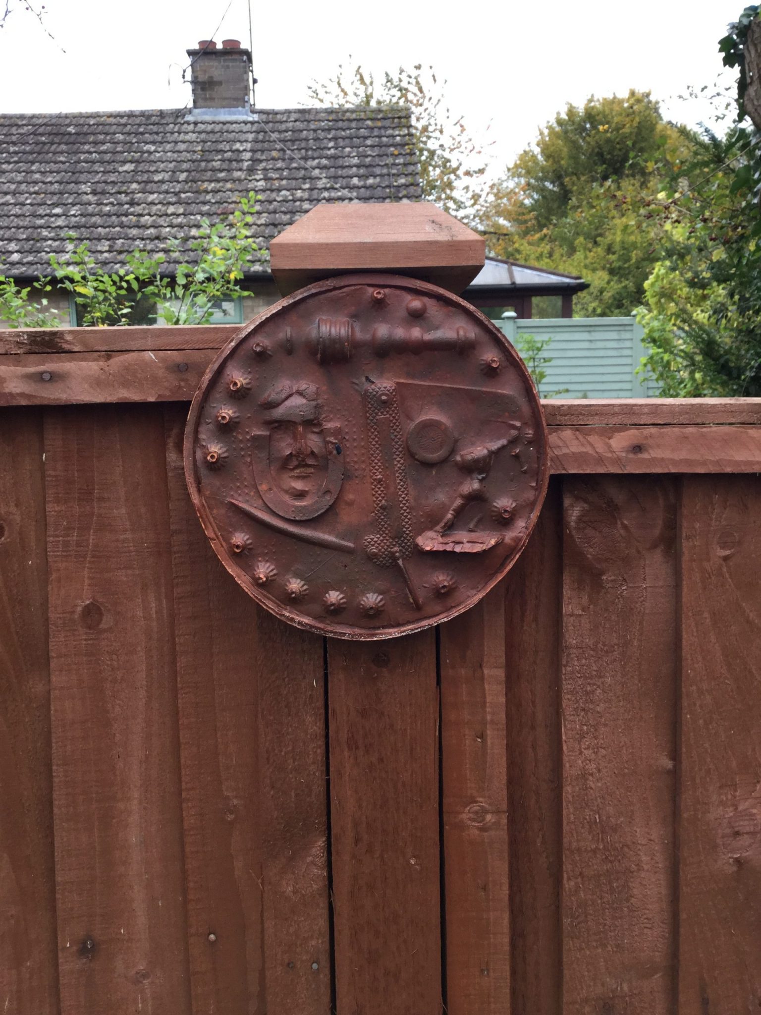 A brown circular plaque with impressions of a small horse shoe, a toy and a face, on a garden fence