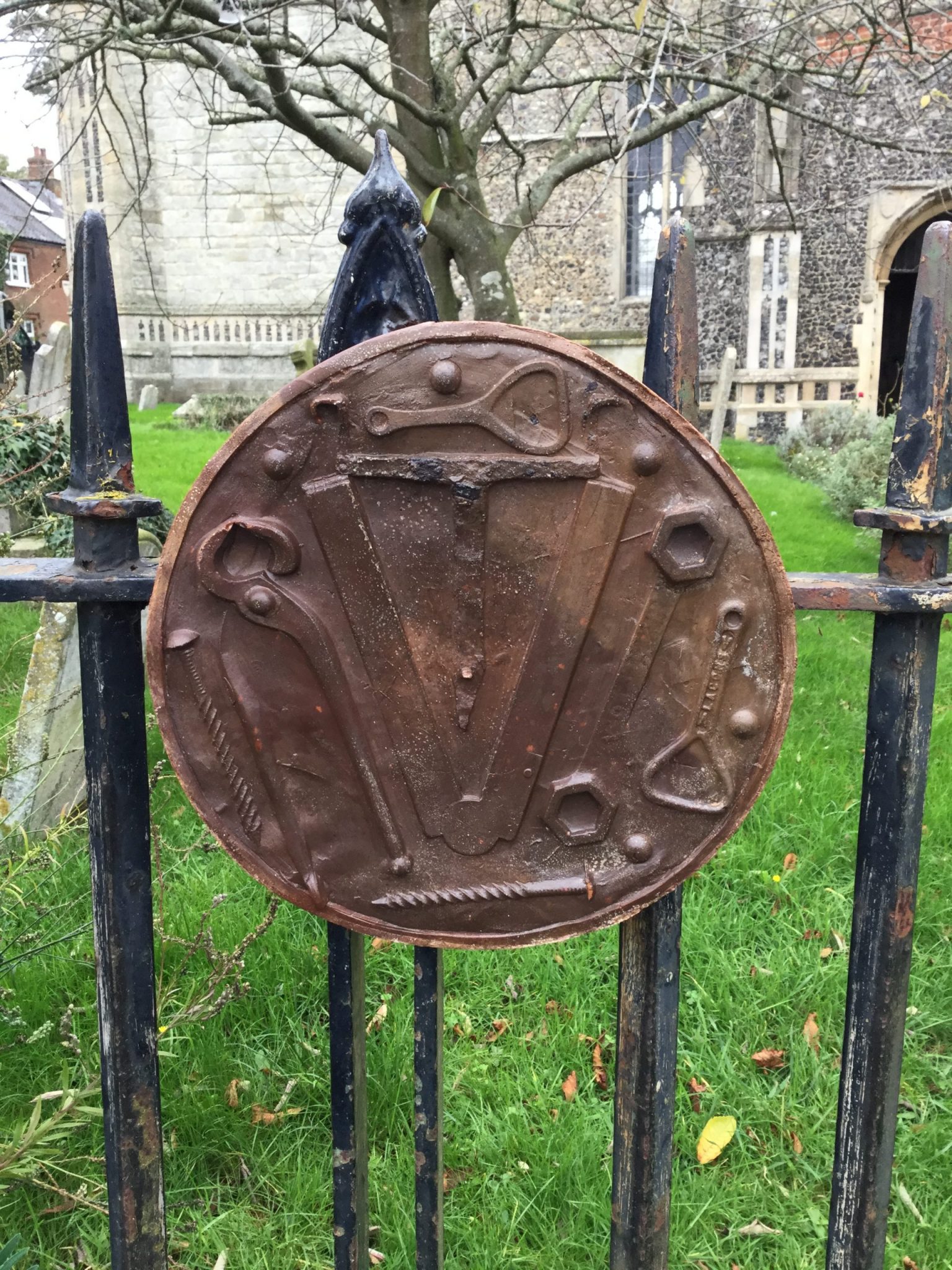 A brown circular plaque with impressions of pincers, a folded rule and a spanner, on the church railings