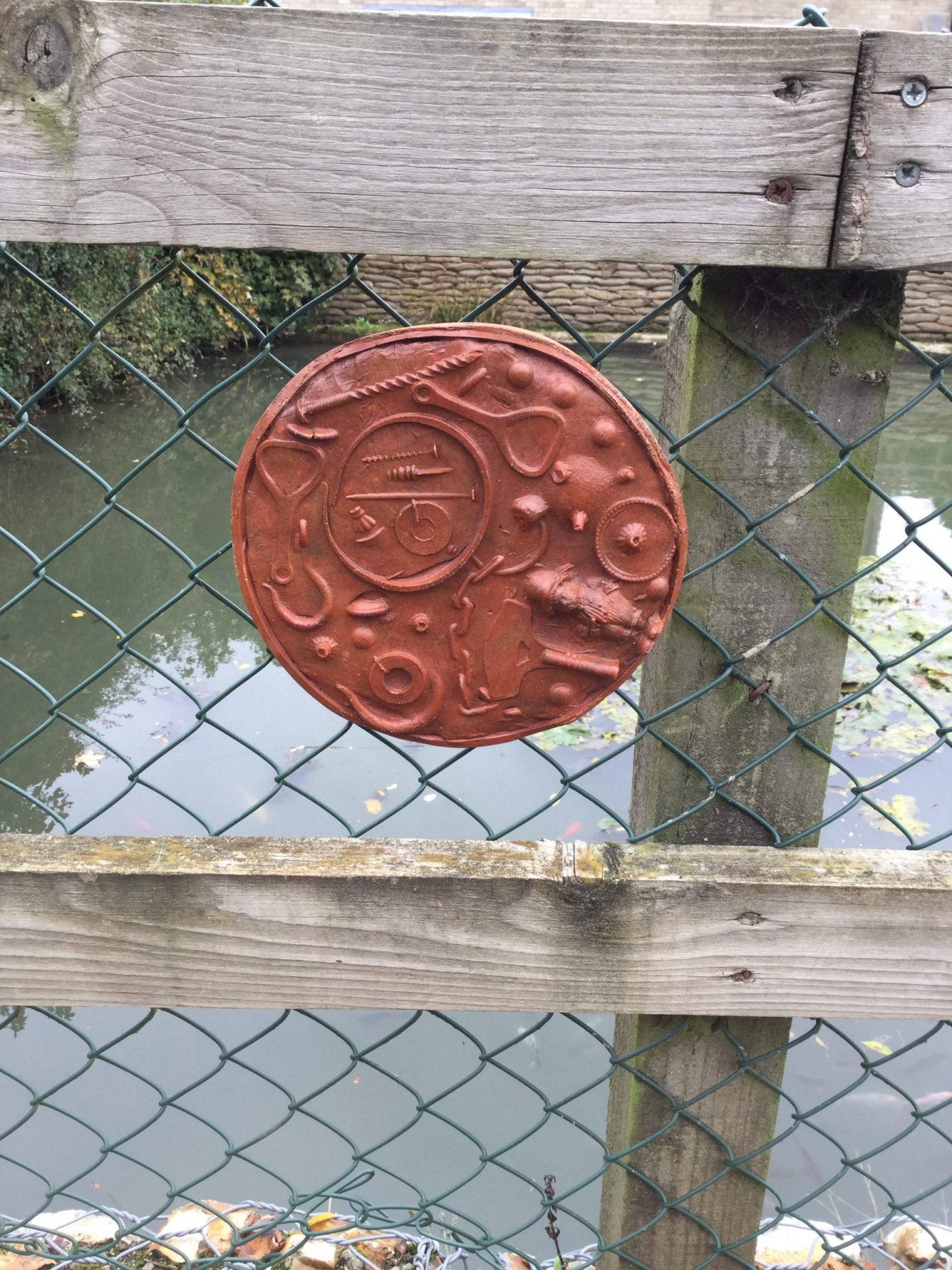 A circular plaque, with impression of objects including a bottle opener, hooks and a small toy, attached to a chain link fence in front of the village pond