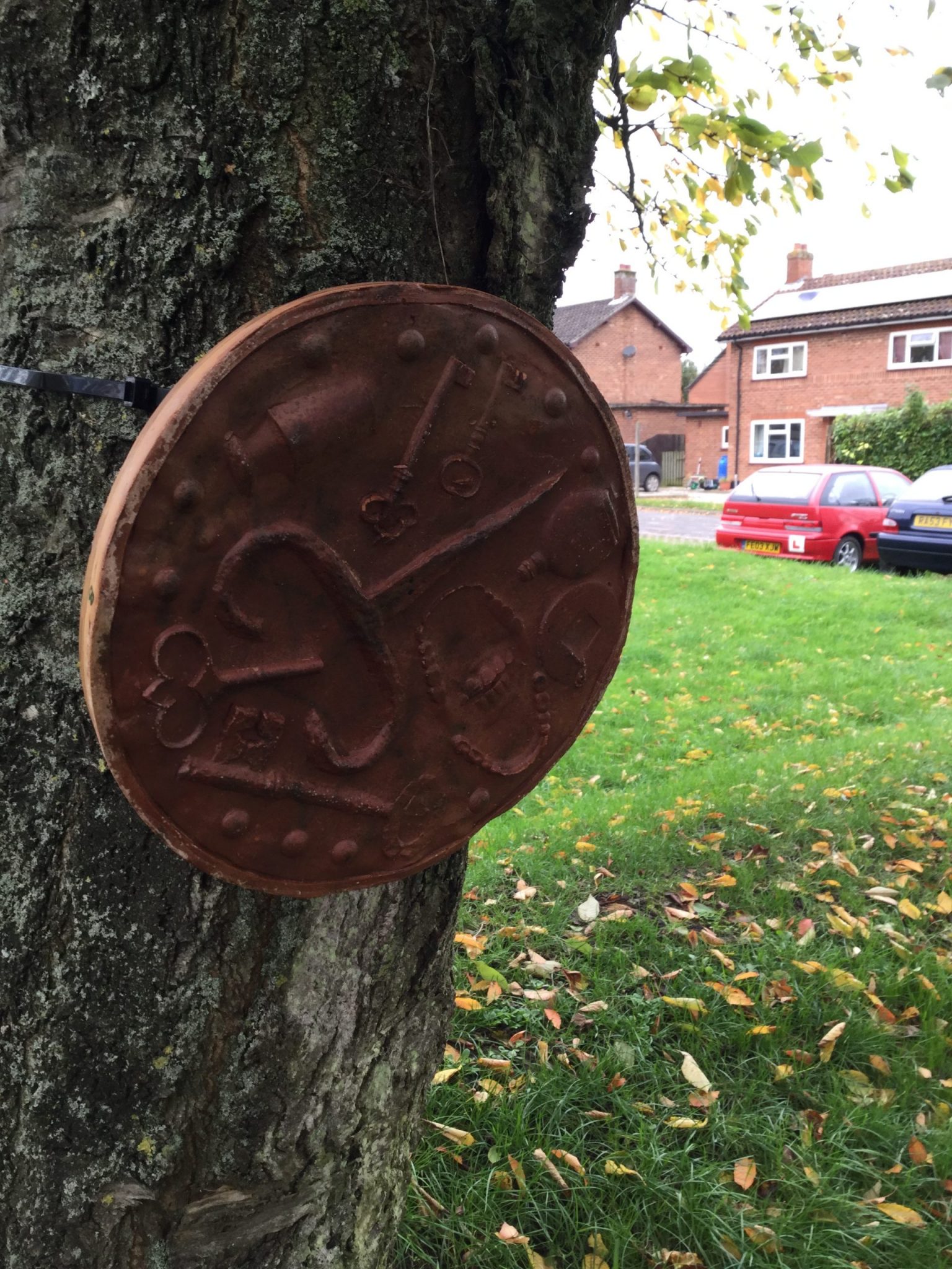 A brown circular plaque with impressions of keys, a small bottle and a pair of false teeth, attached to a tree