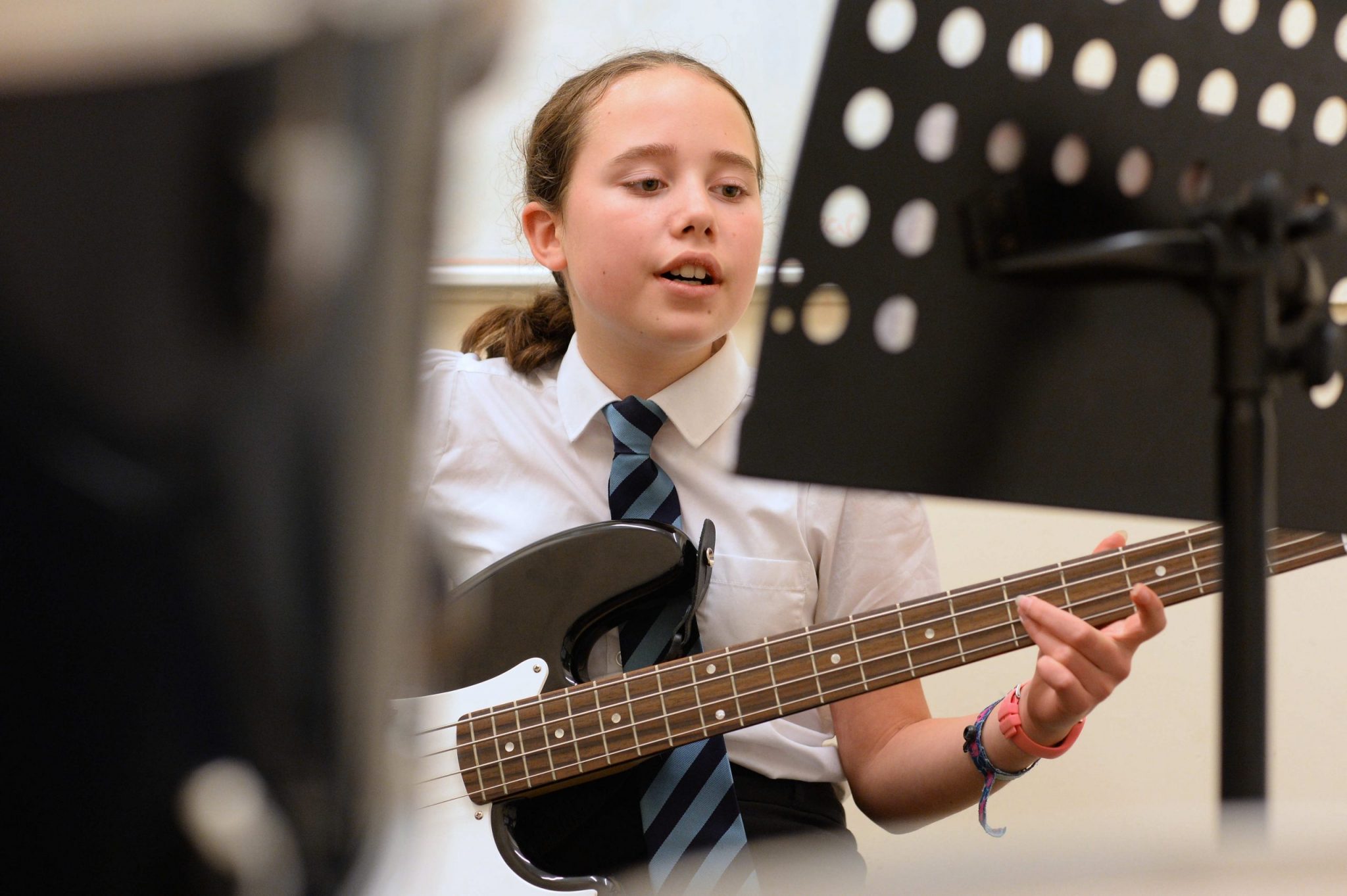 A young girl is playing bass guitar whilst singing from a sheet on a music stand in front of her