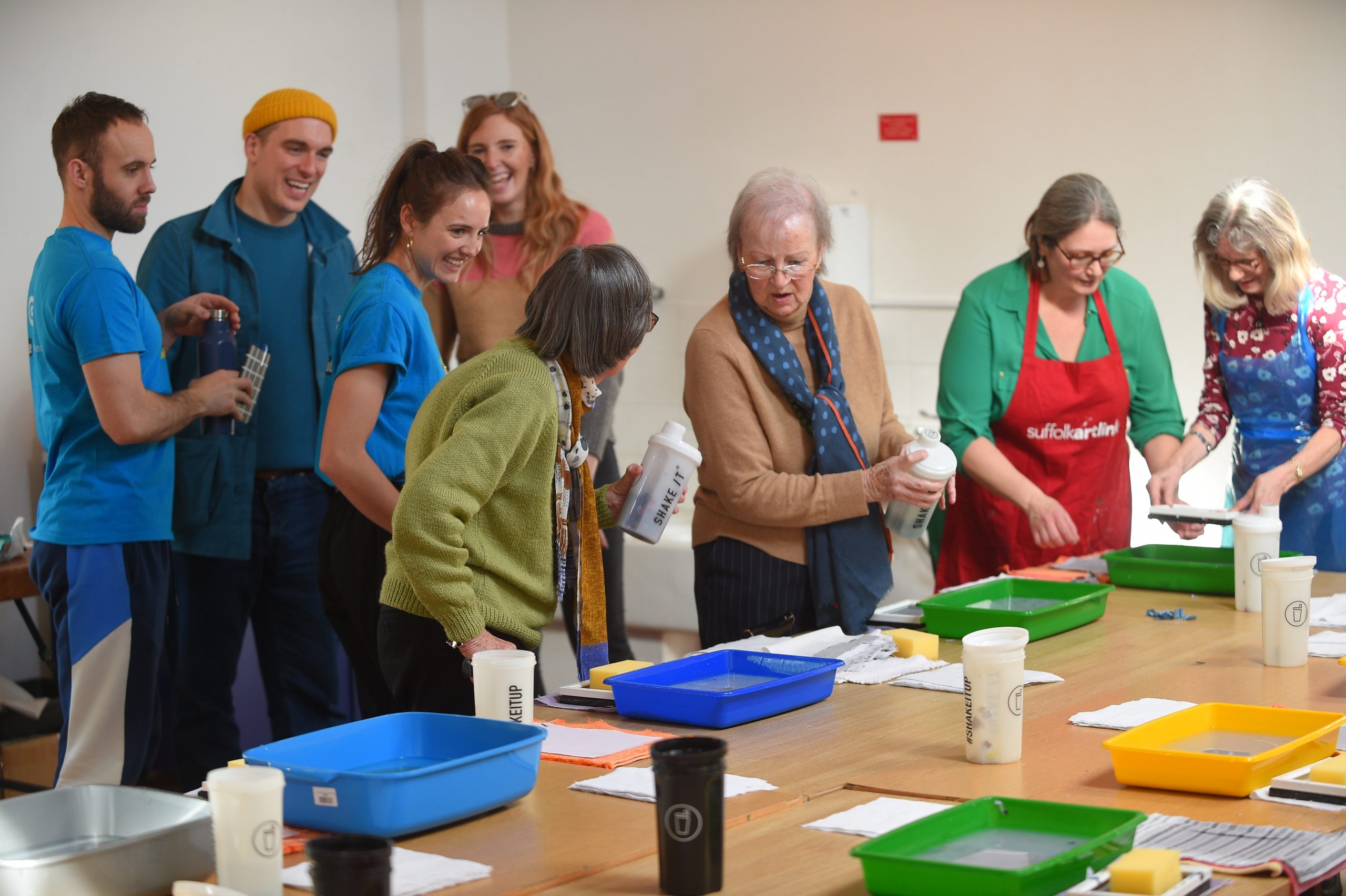 A group of people around a large table, laid out with paper making materials. One woman holds a flask on which are written the words shake it