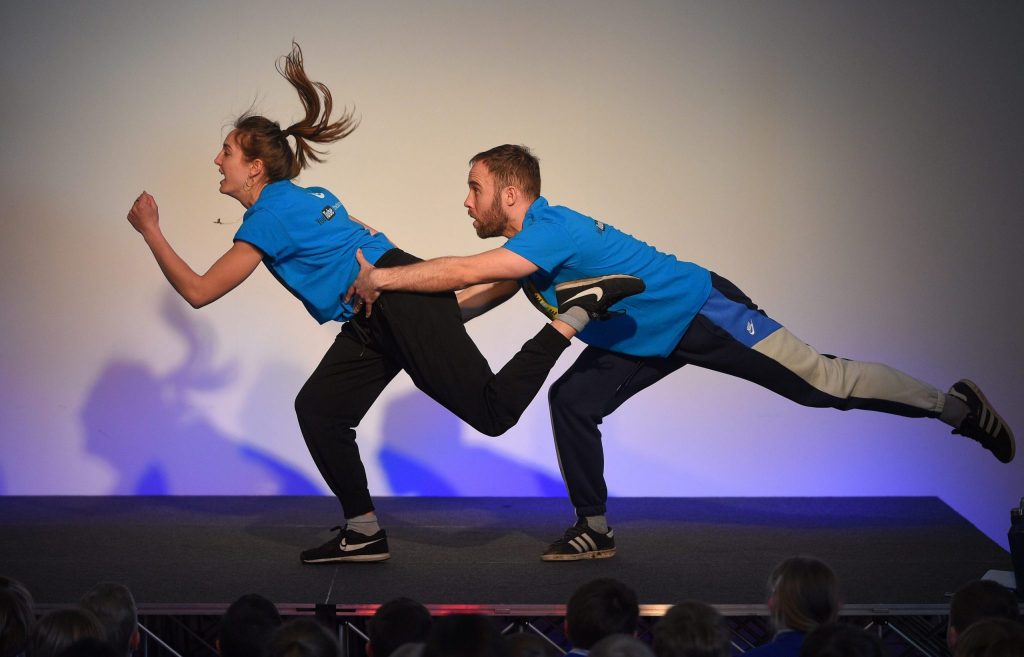 Two dancers, dressed in vivid blue T shirts and black leggings, dancing against a white background