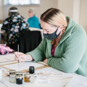 Participant holding paintbrush above page with small pots of paint in the foreground