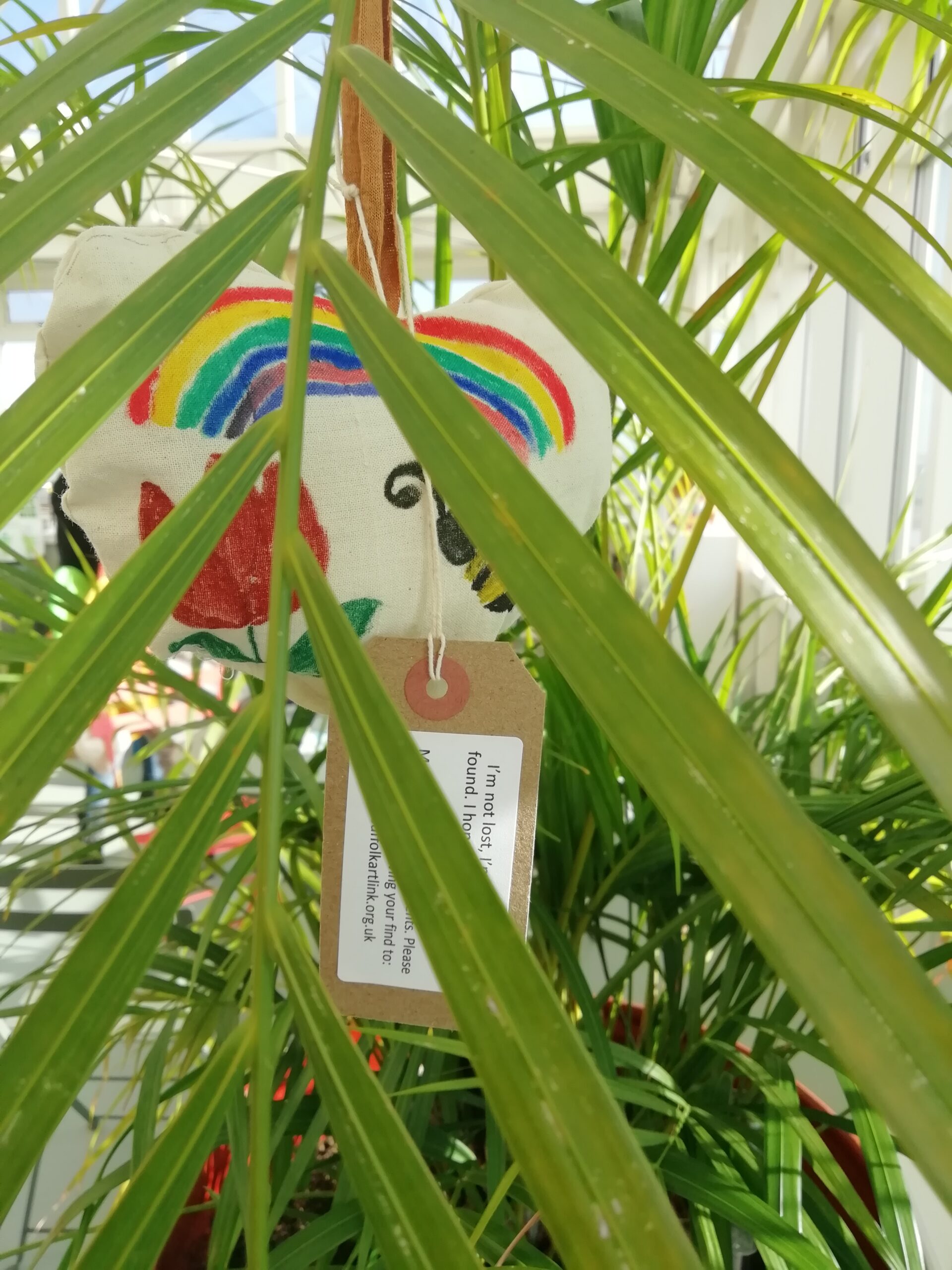 White heart decorated with a bee, a rainbow and a red rainbow hanging in a plant and partially obscured by foliage