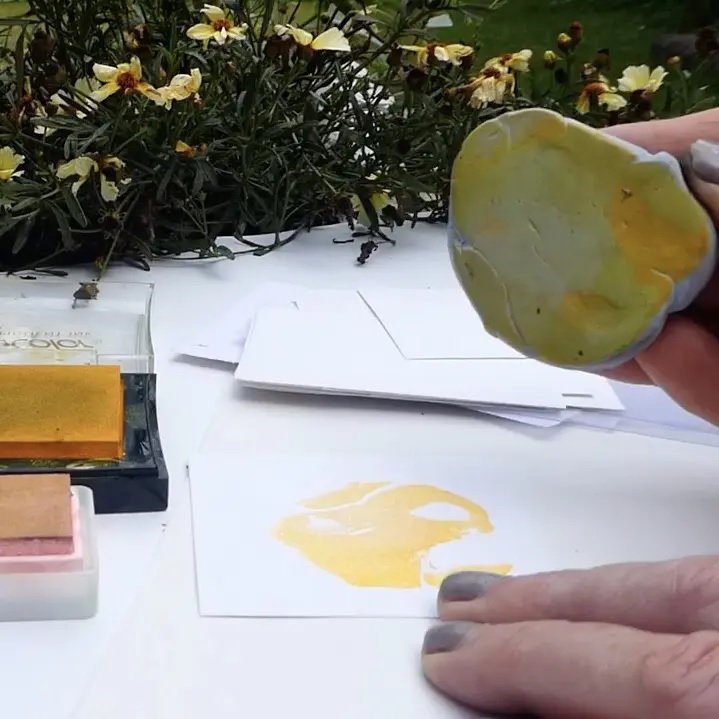 A hand holding a piece of yellow-inked Blutack over a white sheet of paper, having printed a yellow shape onto the paper