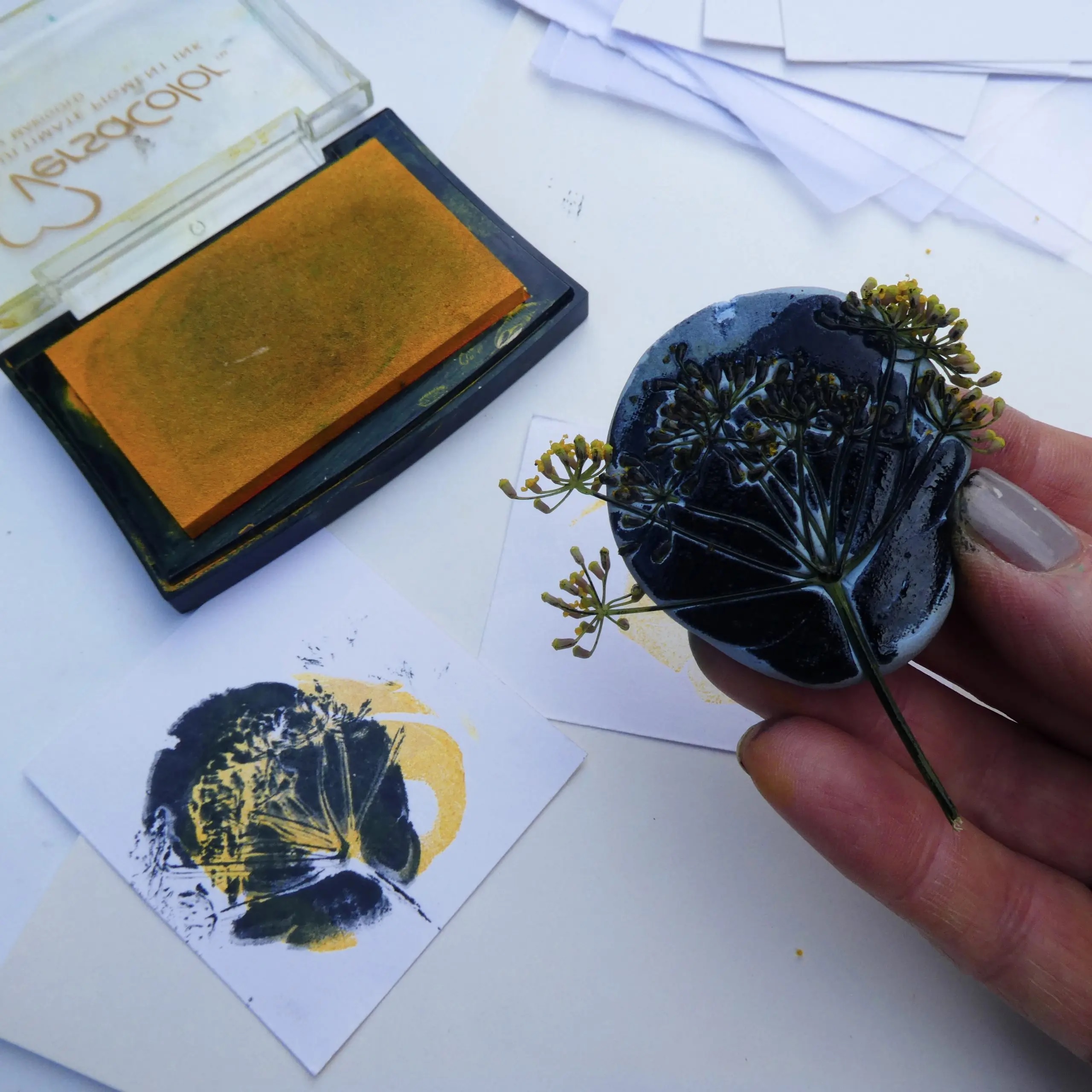 A hand holds an inked up Blutack with seed head and in front is the print made using it
