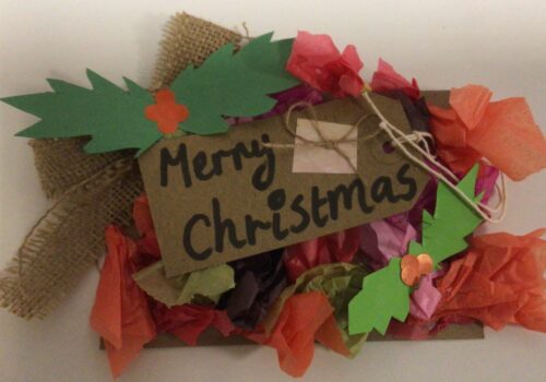 A postcard decorated to look like a Christmas wreath with paper holly leaves and tissue paper