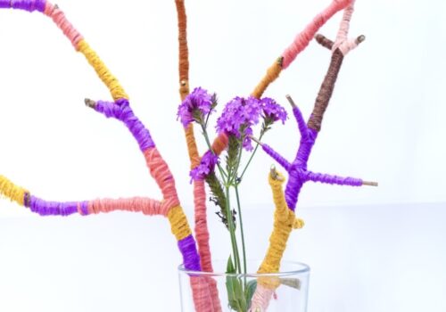 Yarn wrapped stick and purple flower in a glass. Stick wrapped in yellow, peach, orange, brown and purple yarn.