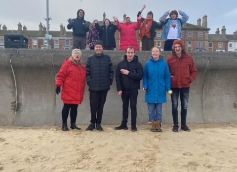 11 students and staff on Lowestoft Beach waving their hands to the camera. It was a cold day so everyone is dressed in warm coats and hats.