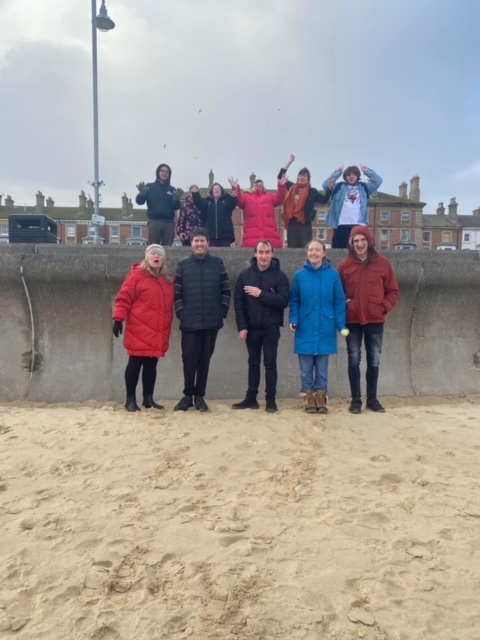 11 students and staff on Lowestoft Beach waving their hands to the camera. It was a cold day so everyone is dressed in warm coats and hats.