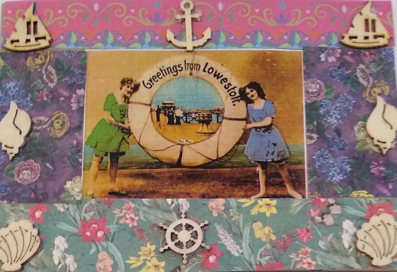 A postcard collaged with a background of strips of patterned paper in pinks, blues and greens and featuring a central image of a vintage postcard bearing the message Greetings from Lowestoft and surrounded by tiny wooden cut out shapes of boats, shells, a ship's wheel and an anchor