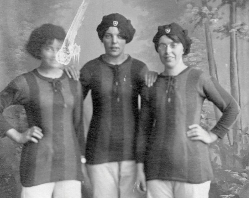 An old monotone photograph of three women stand side by side, they are dressed in vertically striped long sleeved shirts, light coloured shorts and wearing bonnet-like caps on their heads.