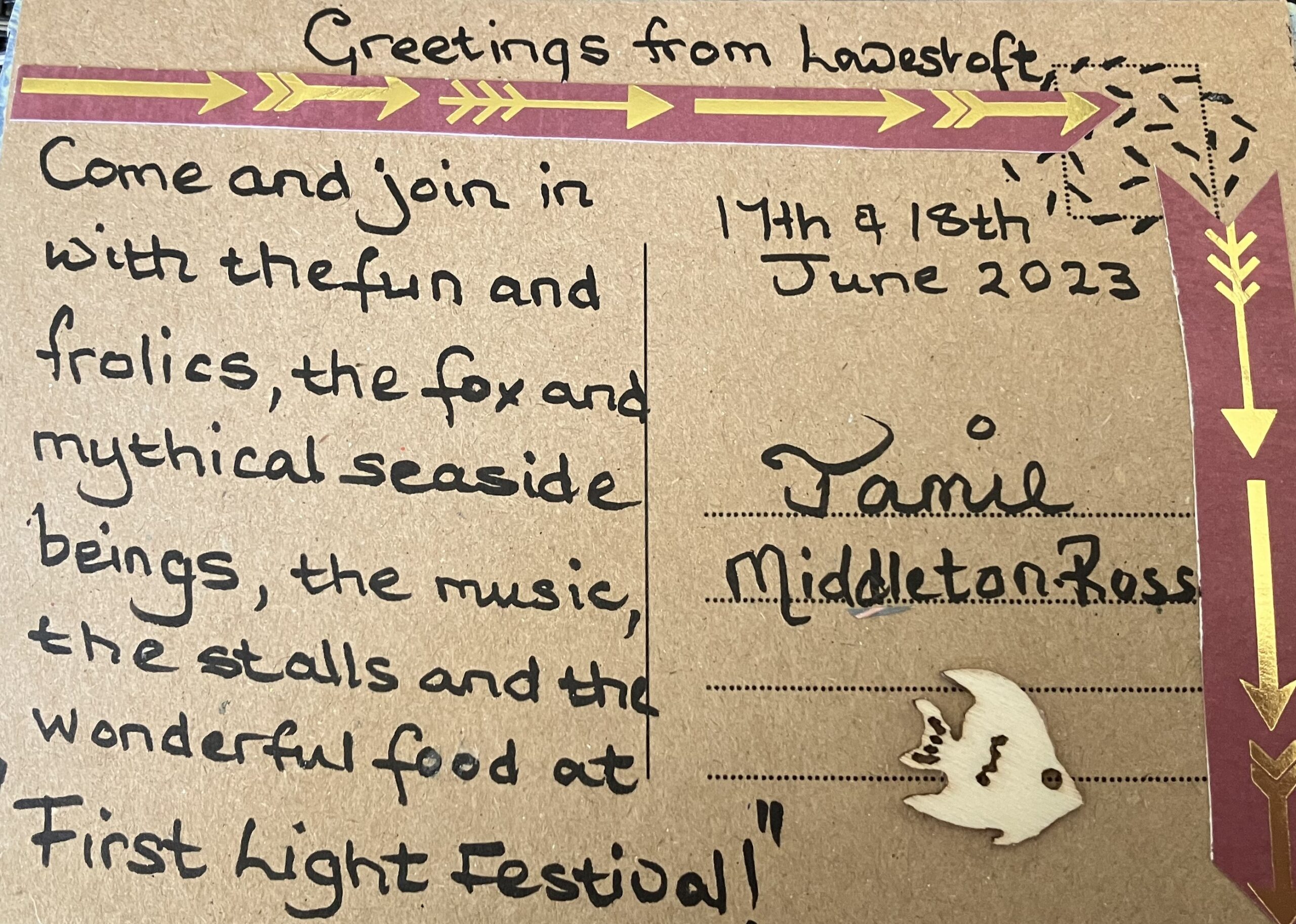 Postcard decorated with gold arrowed tape and small wooden fish cut out with text reading Greetings from Lowestoft Come and join in with the fun and frolics, the fox and mythical seaside beings, the music, the stall and the wonderful food at First Light Festival 17th and 18th June 2023