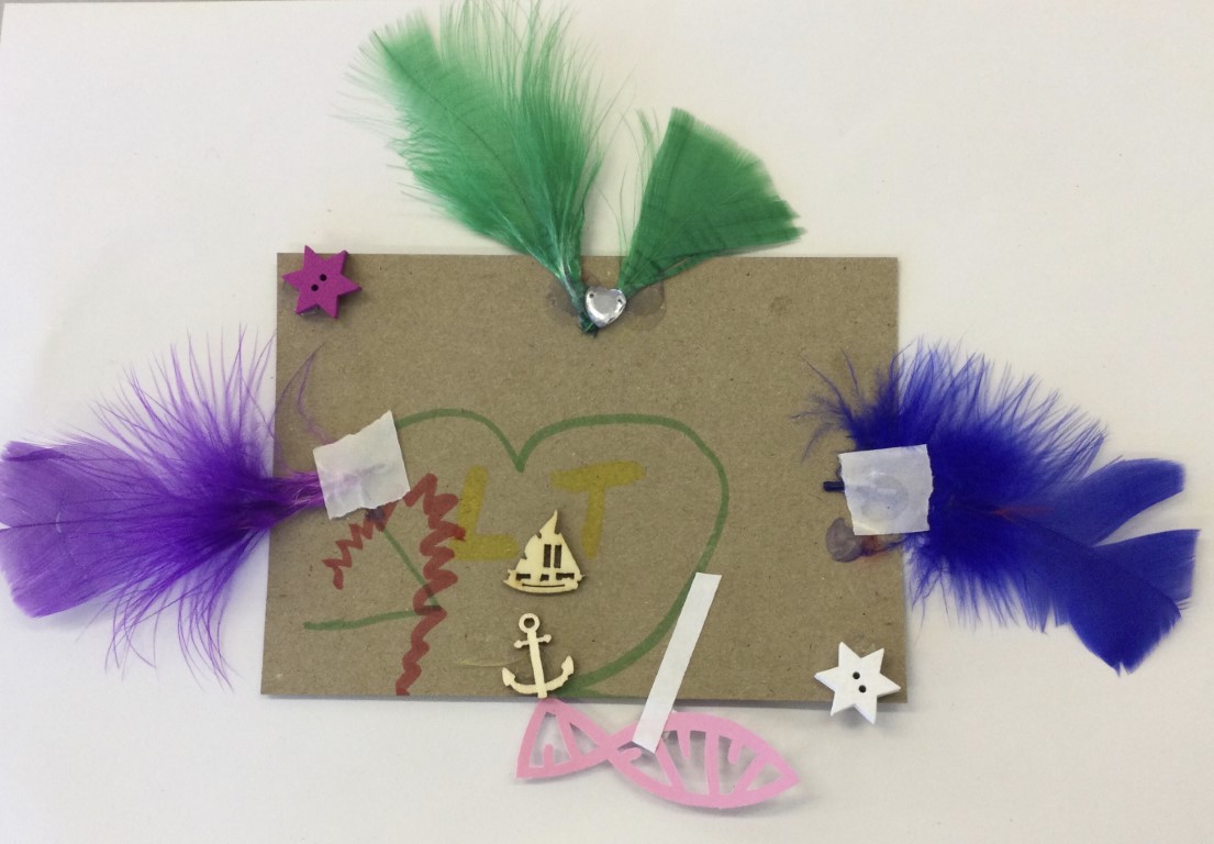 This brown postcard has a hand-drawn heart, drawn in green felt tip pen, inside which are glued two small shapes - one of a boat and the other an anchor. A single purple feather is glued on the left hand side of the card and a blue one is on the right. At the top of the card, 2 green feathers create a V shape in the middle of which is a small shiny heart. A pink fish is attached by a piece of tape so that it appears to be swimming below the card and there are also two wooden stars, one white and one purple.