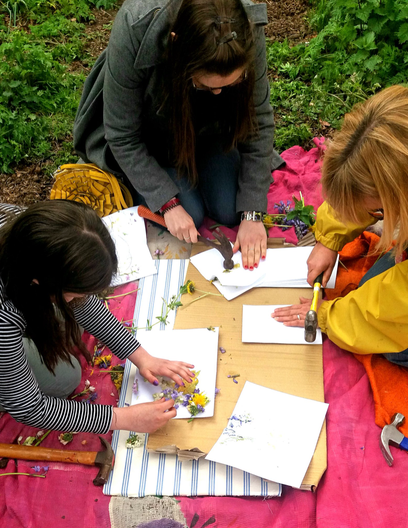 Three people are kneeling on the ground on a pink blanket on which is some blue and white striped fabric and cardboard on top of that. Each person has some sheets of white paper in front of them; one person is arranging flowers on one half of a piece of paper and the other two have folded over their papers and are using hammers to flatten the plants beneath the top sheet