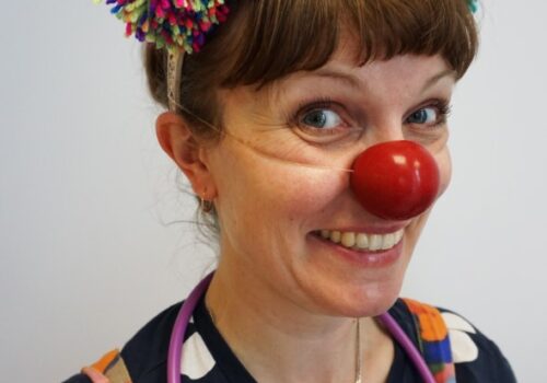 Dr. Dot-to-Dots, Clown Doctor, wearing a hat of colourful pompoms and pencils, and a toy stethoscope around her neck.