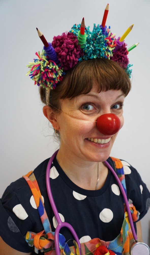 Dr. Dot-to-Dots, Clown Doctor, wearing a hat of colourful pompoms and pencils, and a toy stethoscope around her neck.