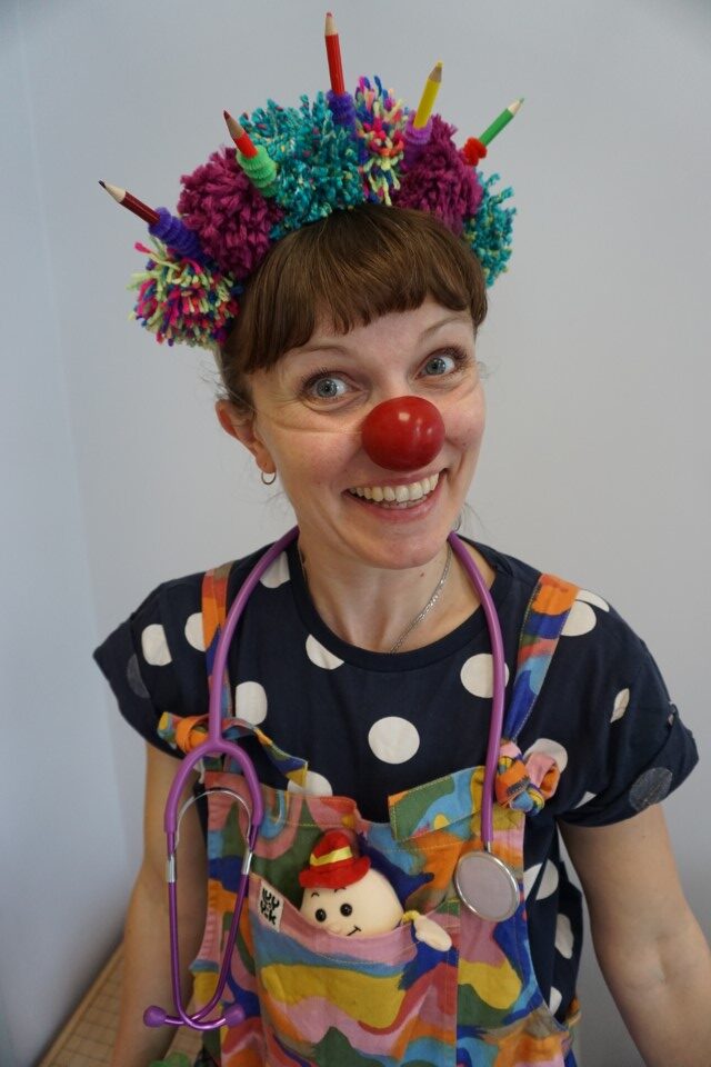 Dr. Dot-to-Dots, Clown Doctor, wearing a hat of colourful pompoms and pencils, and a toy stethoscope around her neck.