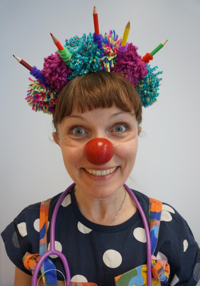 Dr. Dot-to-Dots, Clown Doctor, wearing a hat of colourful pompoms and pencils, and a toy stethoscope around her neck.