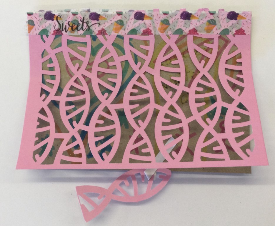 A brown postcard decorated in different coloured felt tip pens, over which is laid a sheet of pink card with a cutaway pattern resembling fishes. Along the top is a decorated tape with the word Sweets on it and, suspended from the bottom of the card is a pink fish.
