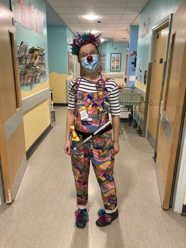Dr. Dot-to-Dots, Clown Doctor, wearing a hat of colourful pompoms and pencils, and a toy stethoscope around her neck. Dr. Dot-to-Dots is making her ward visits.