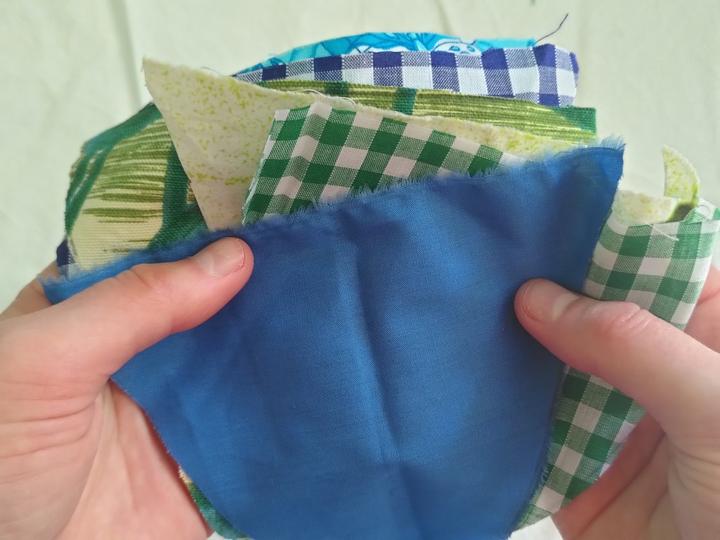 A pair of hands holds a selection of fabric bunting flags, the top one is plain blue and underneath can be seen edges of green gingham and blue gingham flags, as well as other patterned materials.