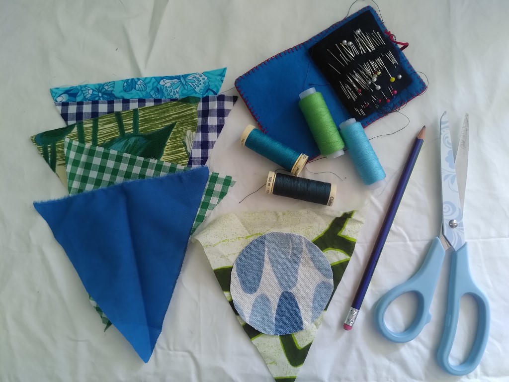 A selection of bunting flags in different coloured and patterned materials is laid out on a white cloth background beside four spools of cotton, a blue hand-stitched pin case holding lots of pins and needles, a blue pencil and a pair of scissors. Centre is a patterned bunting flag overlaid with a circle of blue patterned fabric