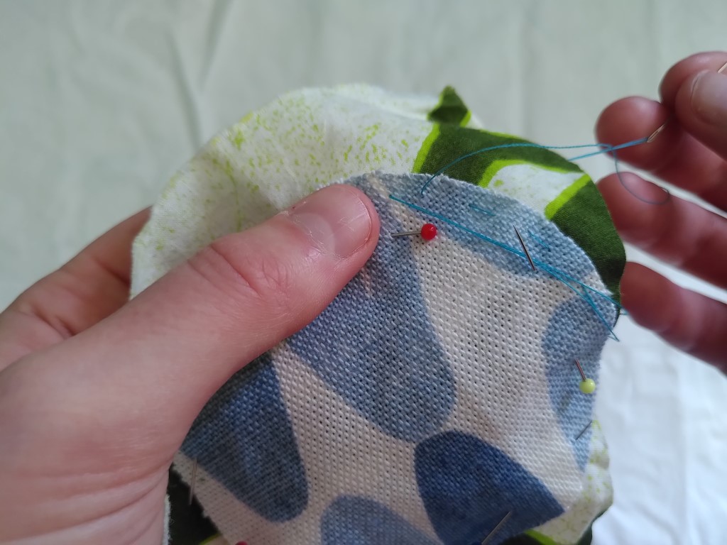 Close up of a hand holding several layers of fabric that are pinned together. The other hand holds a needle thread with blue cotton which they are using to stitch the pieces of fabric together.