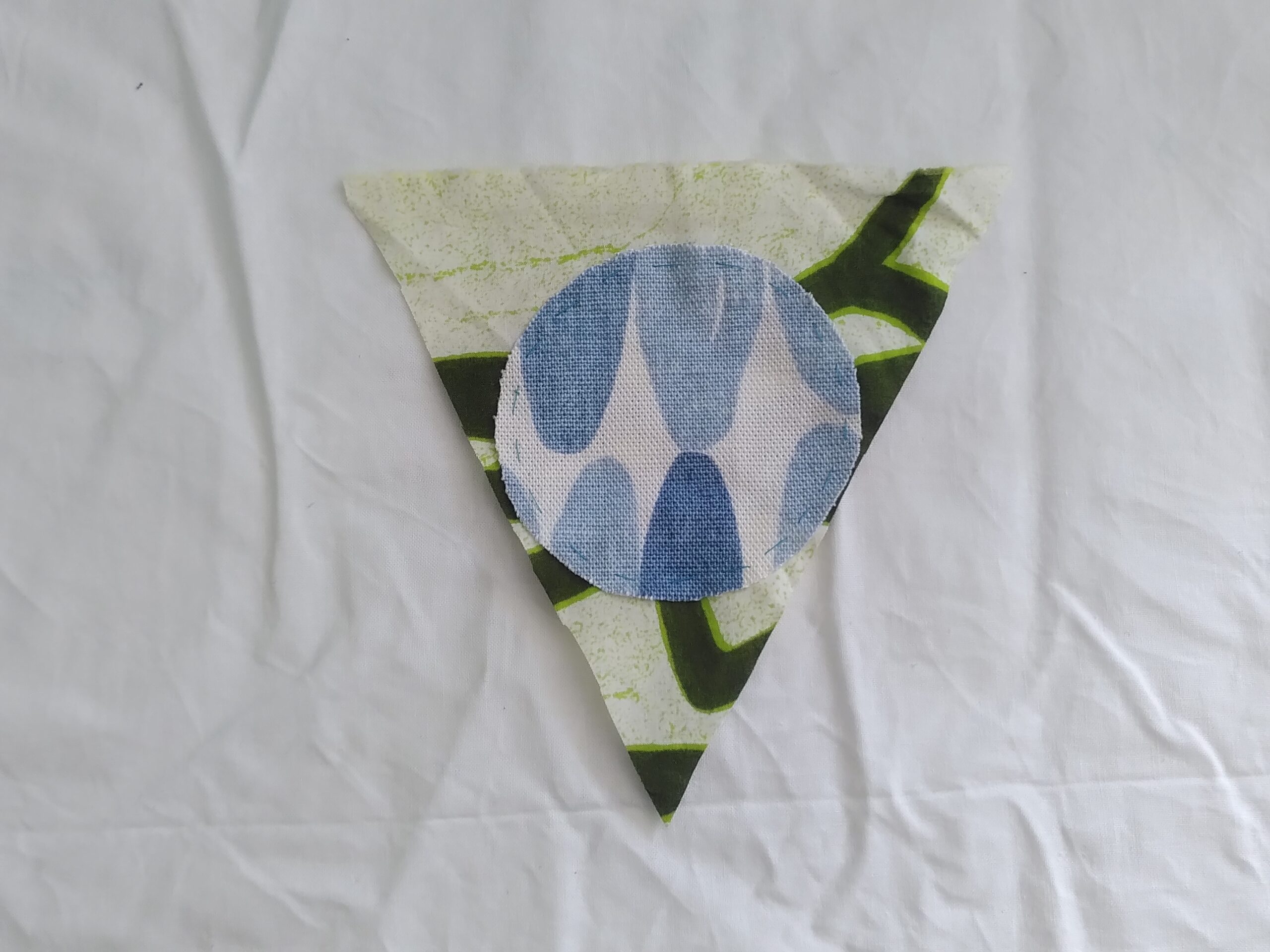 On a creased white fabric background lies a triangle of material, patterned with thick green lines over which is placed a small circle of fabric with blue patterns on it