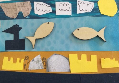 A postcard collaged with paper and wooden shapes, with a black border along the bottom, above which are cut-out shapes of sandcastles, stones and hand drawn people. Above it a strip of blue paper with two wooden fish glued and a black paper boat. The sky above is created with a strip of blue patterned paper, overlaid with white clouds and a yellow sun